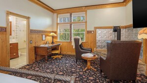 A room with a wooden desk, a chair, a small table, and a window with a view of greenery.