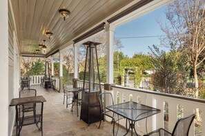 A covered outdoor patio with tables and chairs, a tall metal heater, and a view of trees and a fence.