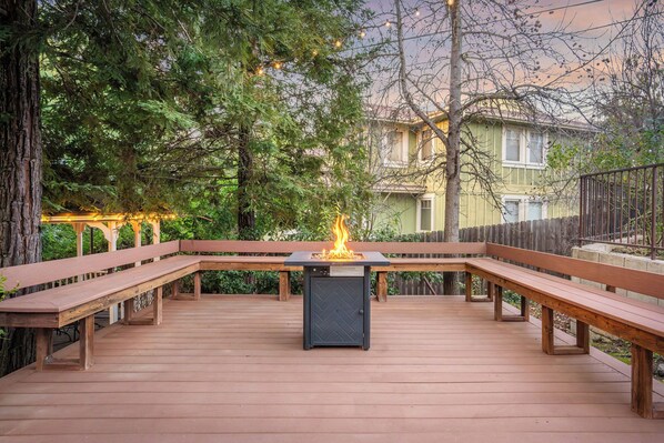 An outdoor deck with a built-in fire feature, wooden benches, and a view of a house in the background.