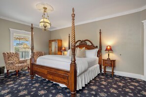 A four-poster bed with a wooden frame and white bedding, two bedside tables with lamps, a floral-patterned armchair, and a view of the outdoors through a window. - Demetria by Polo House and Bungalows (Santa Ynez)