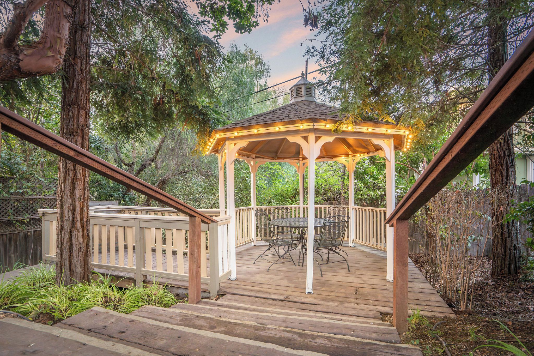 A gazebo with a wooden deck, surrounded by trees and a wooden fence.