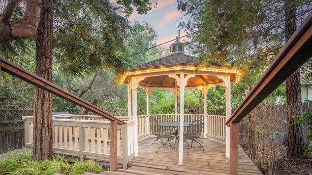A gazebo with a wooden deck, surrounded by trees and a wooden fence.