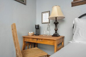 Un dormitorio ordenado con una mesita de noche de madera, una cama con ropa blanca, una lámpara, una cafetera y un certificado enmarcado en la pared.