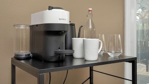 A Nespresso coffee machine on a table with two white cups, a glass, and a bottle.