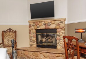 Ein Wohnzimmer mit Kamin aus Stein, einem an der Wand befestigten Fernseher, einem Holzstuhl und einem Schreibtisch mit Lampe.