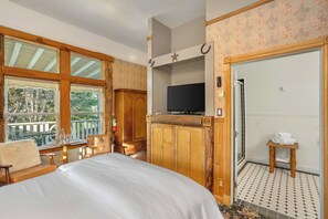 A bedroom with a large bed, wooden wardrobe, a television, and a balcony with a view of trees.