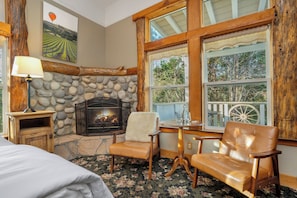 A cozy room with a stone fireplace, wooden furniture, and large windows offering views of trees.