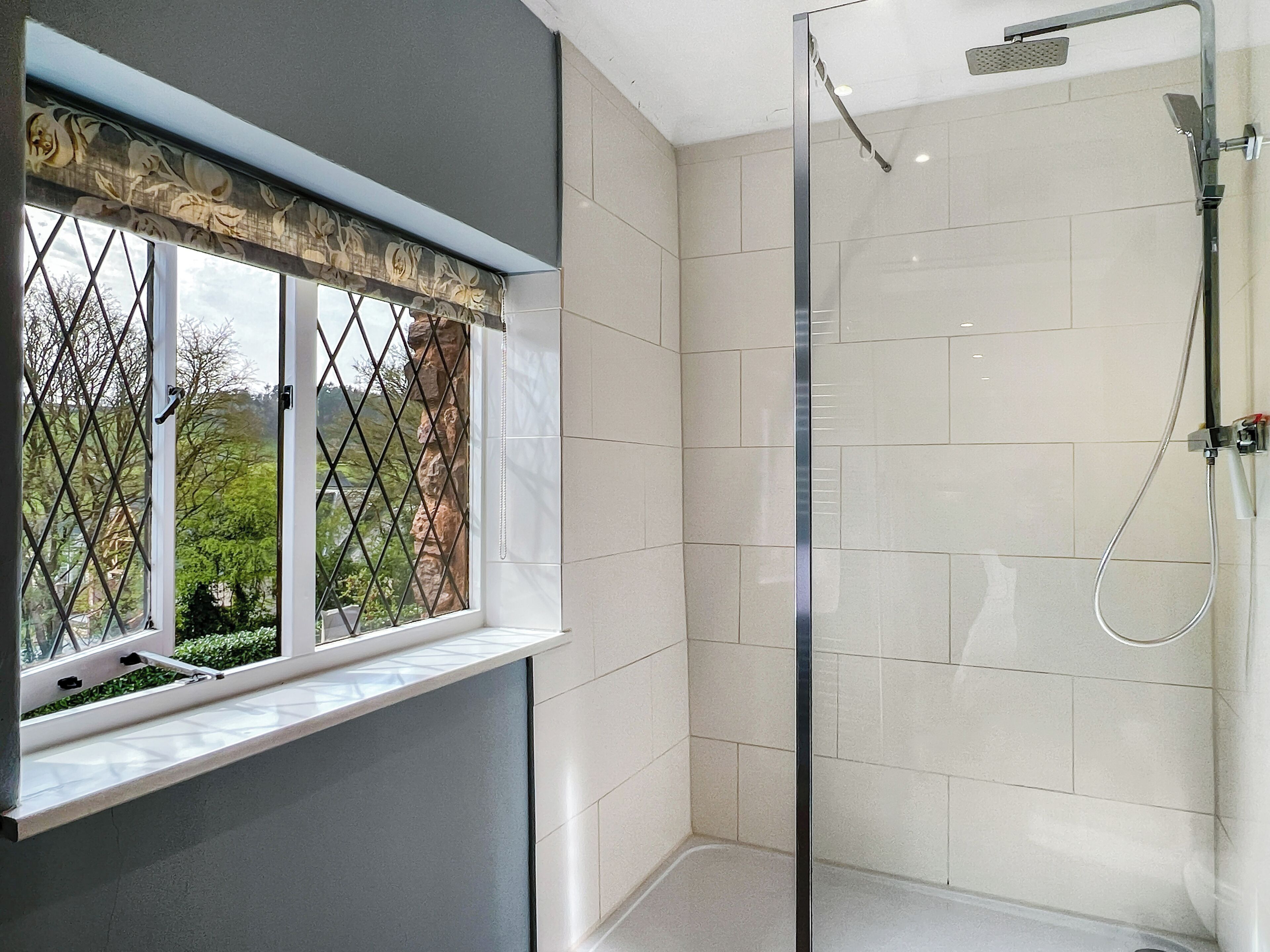 Un baño moderno con cabina de ducha de vidrio, una ventana con un adornado valance y vistas a la vegetación.