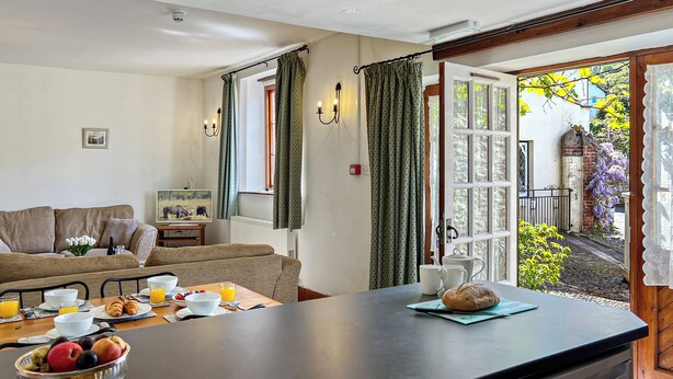A cozy living room with a sofa, a dining table set with breakfast, and a view of a garden through an open door.