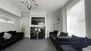 A living room with two black sofas, a fireplace with a vase of flowers, a chandelier, and a large window with blinds. - Watton By Verve (Thetford)