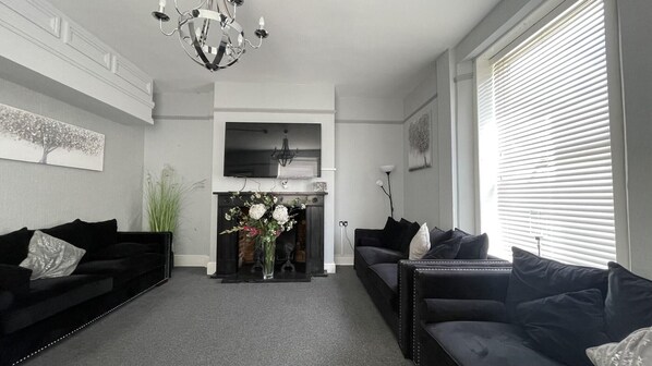 Uma sala de estar com dois sofás pretos, uma lareira com arranjo floral, um candelabro e uma janela grande com persianas.