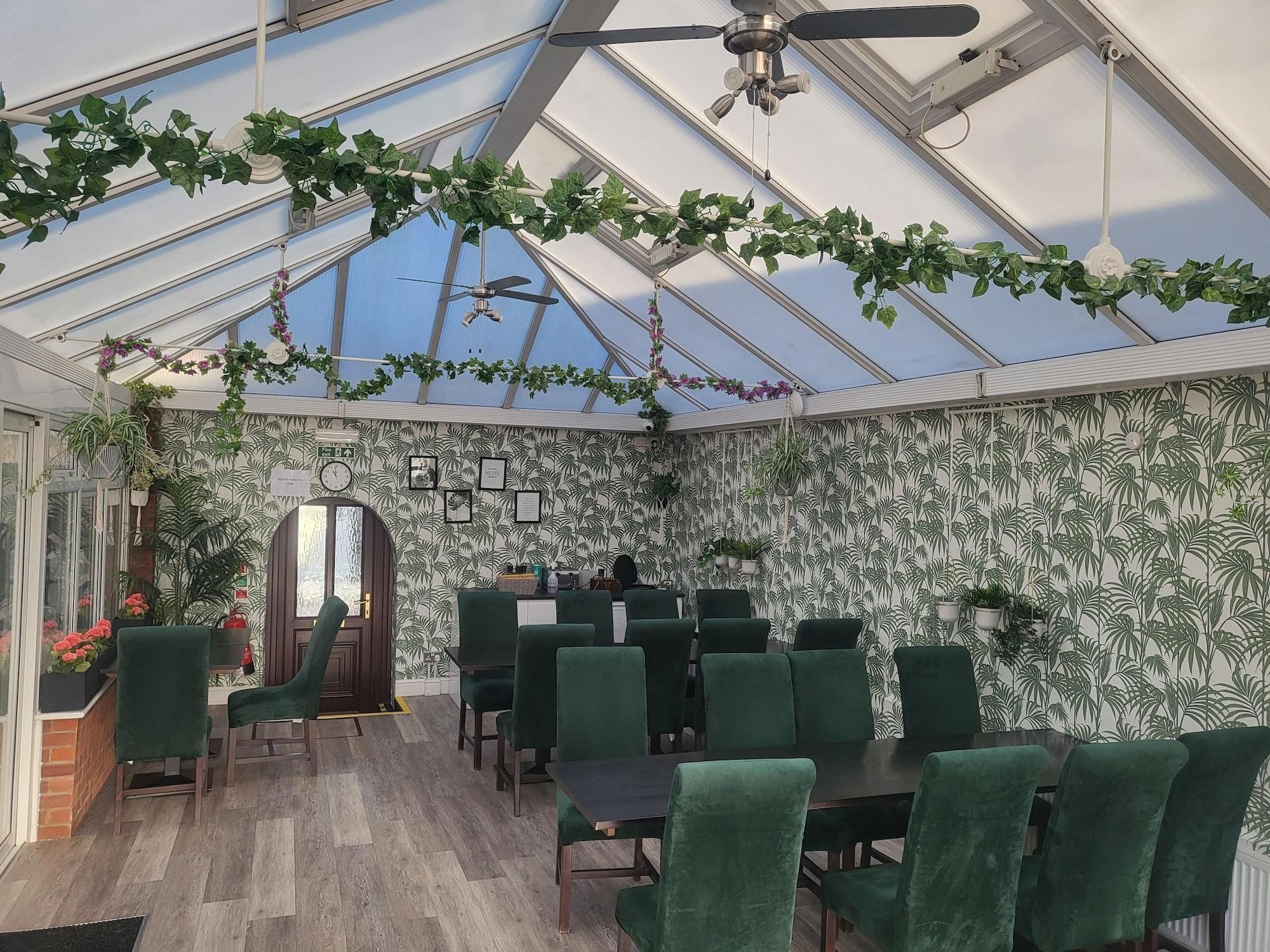 A conservatory with a glass ceiling, green chairs, and potted plants.