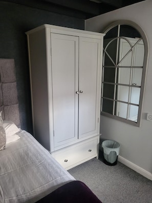 Un dormitorio con cama, armario blanco, un cesto de basura pequeño y una ventana de arco.