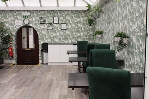 A room with a wooden door, a fire exit sign, a clock, framed pictures on the wall, and green upholstered chairs. - Watton By Verve (Thetford)