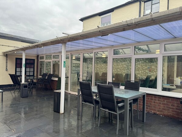 A covered outdoor seating area with tables and chairs, large windows, and a brick wall. - Watton By Verve (Thetford)