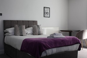 A modern bedroom with a grey upholstered bed, white bedding, and a purple throw blanket. A motivational quote is displayed on the wall. - Watton By Verve (Thetford)