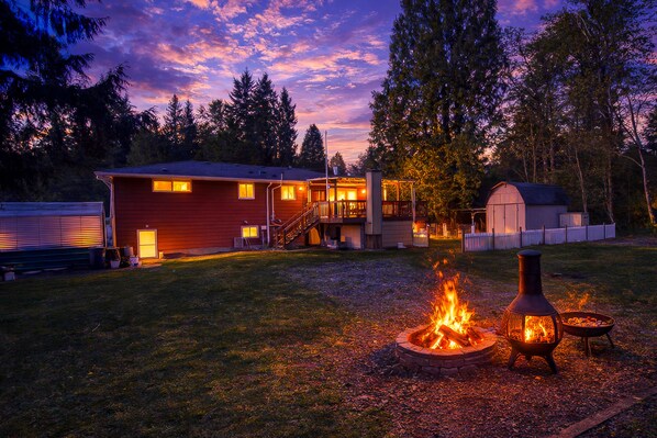 A rustic cabin with a fire pit and a lantern, surrounded by a wooden deck and a fenced yard.