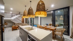 A modern kitchen with a central island, a dining area with wicker chairs, and a staircase leading to an upper level. - Kammara 1 Villa Bali (Canggu)