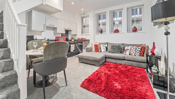 Ein modernes Wohnzimmer mit einem grauen Sofa, einem roten Teppich und einem Essbereich mit Tisch und Stühlen.