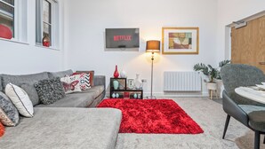 Ein modernes Wohnzimmer mit einem grauen Sofa, einem roten Teppich, einem schwarzen Beistelltisch, einer Lampe und einem an der Wand montierten Fernseher, der 'NETFLIX' zeigt.