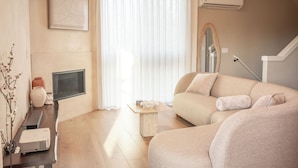 Ein Wohnzimmer mit einer Couch, einem Flachbildfernseher, einem Projektor und einem großen Fenster mit durchsichtigen Vorhängen.
