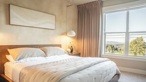 Ein Schlafzimmer mit einem Bett, einem Nachttisch mit Lampe, einem Fenster mit Vorhängen und einem an der Wand hängenden Kunstwerk.