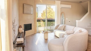 Ein modernes Wohnzimmer mit einer Couch, einem Couchtisch, einem Flachbildfernseher und einem Balkon mit Blick auf Bäume und Blumen.