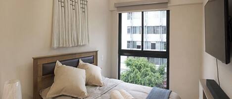 Un dormitorio con cama, ventana, televisor y una obra de arte colgada en la pared.