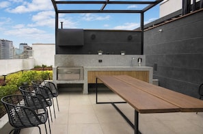 An outdoor kitchen with a built-in grill, a wooden table, and black chairs. - Departamentos Siena (Lima)