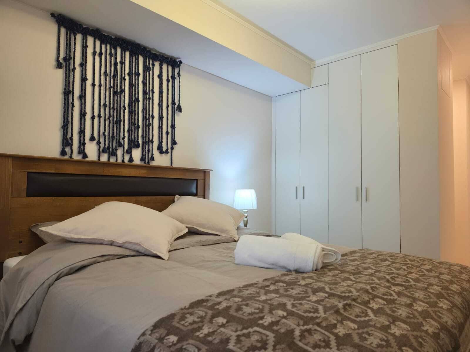 A hotel room with a bed, a headboard, a wall-mounted macrame decoration, a bedside lamp, and a wardrobe.