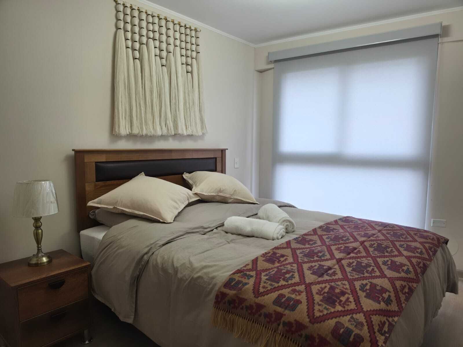 A bedroom with a bed, pillows, a bedside table with a lamp, a patterned blanket, and a window with blinds.