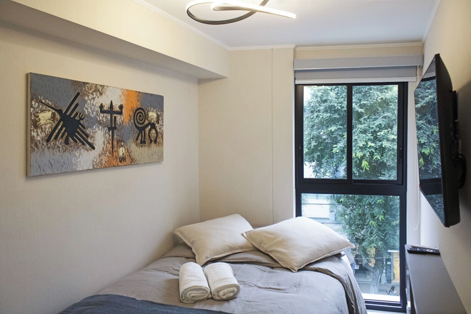 A bedroom with a bed, pillows, a painting, a window, and a TV.