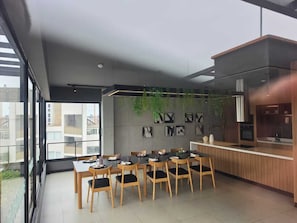A modern dining area with a long table, chairs, and a kitchen area with a counter and appliances. - Departamentos Siena (Lima)
