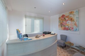 A reception desk with a computer, a printer, and a vase with candles. There is a chair and a glass table with a hat on it. A large floral painting is on the wall. - Lefteris Studios (Naxos)