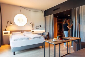 A modern hotel room with a bed, bedside lamps, a table with a cup and a book, and a wardrobe.