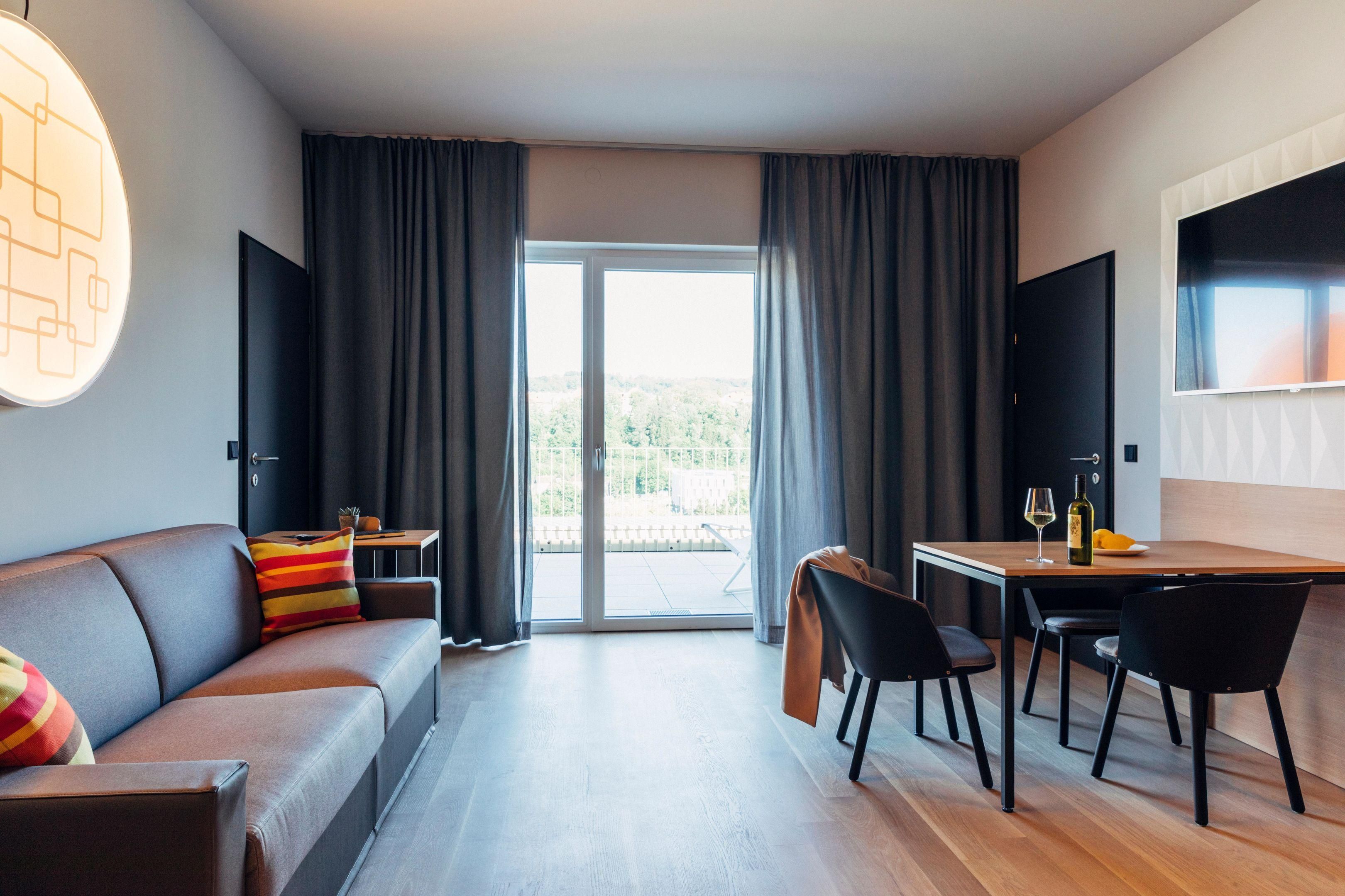 Een moderne hotelkamer met een bank, eettafel, stoelen, een televisie en een glazen schuifdeur die leidt naar een buitenruimte.