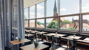 A modern restaurant with large windows, wooden tables, black chairs, and potted plants.