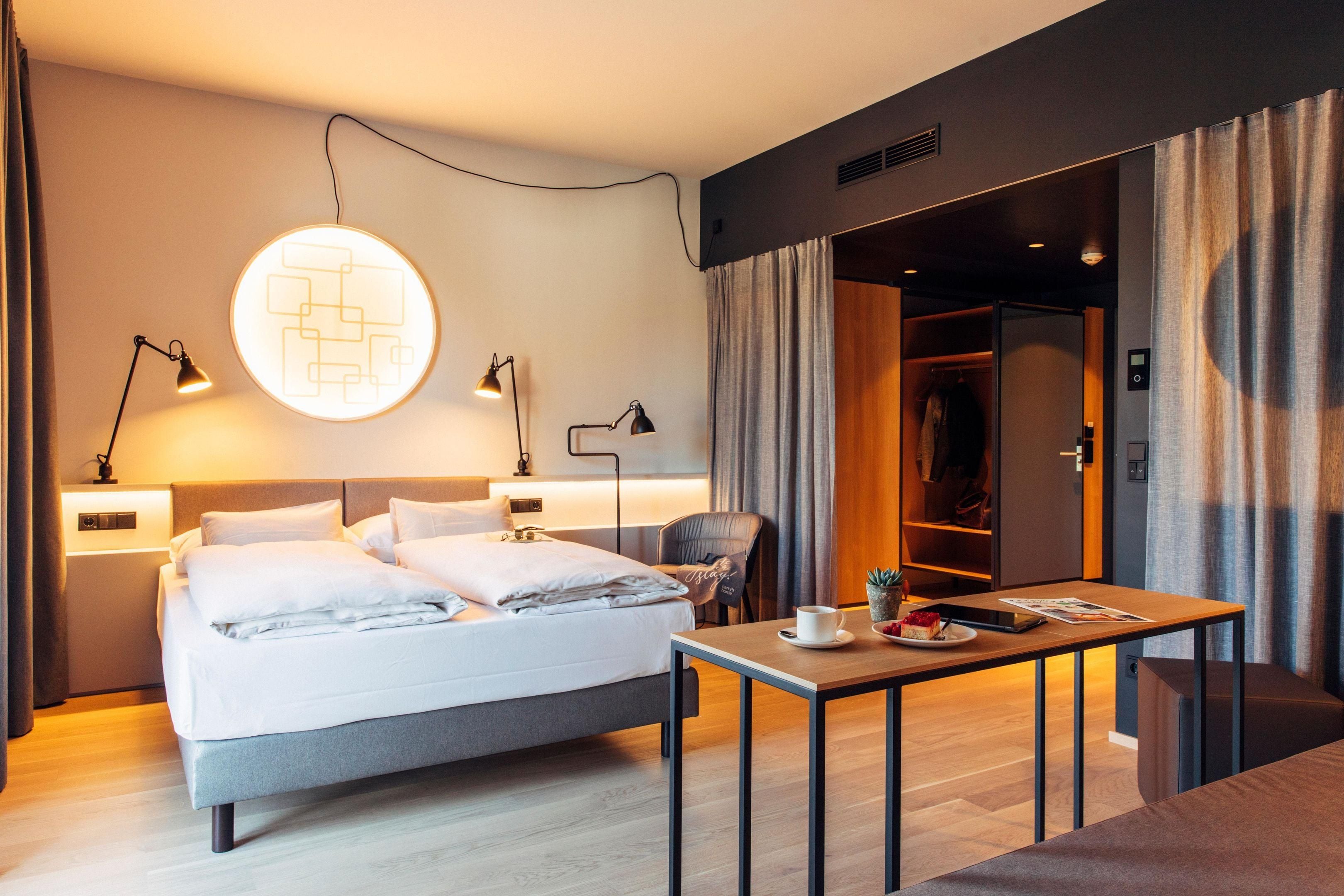 Una camera d'albergo moderna con un letto, lampade ai comodini, un tavolo con una tazza e un libro, e un armadio.