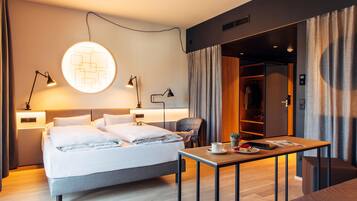 Una camera d'albergo moderna con un letto, lampade ai comodini, un tavolo con una tazza e un libro, e un armadio.