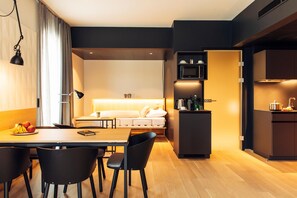 A modern hotel room with a dining area, kitchenette, and a bed with a headboard.