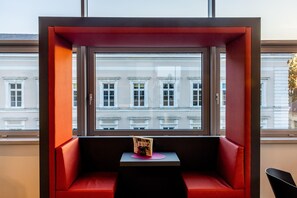 A red cushioned seating area with a small table and a magazine, overlooking a row of white buildings. - Harry's Home Steyr Hotel (Alberndorf in der Riedmark)