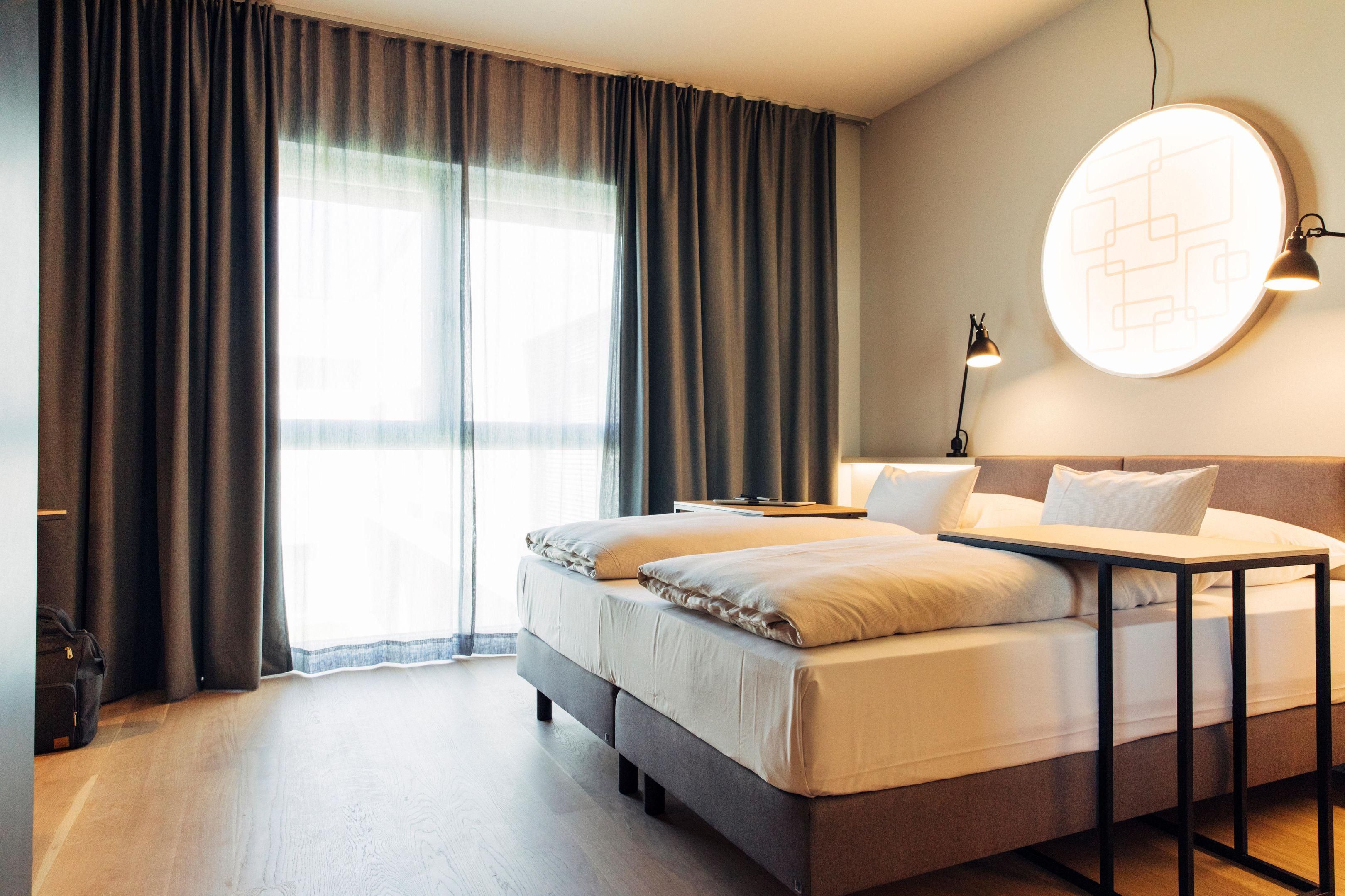 Ein Hotelzimmer mit Bett, Nachttisch, Lampe und einem großen Fenster mit Vorhängen.