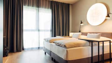 Ein Hotelzimmer mit Bett, Nachttisch, Lampe und einem großen Fenster mit Vorhängen.