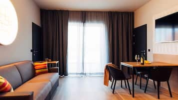 Ein modernes Hotelzimmer mit einer Couch, einem Esstisch, Stühlen, einem Fernseher und einem großen Fenster mit Vorhängen.