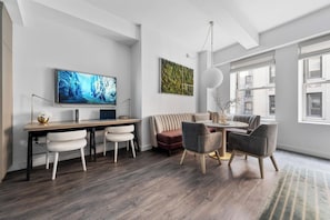 A modern living room with a flat-screen TV, a sofa, a dining table with chairs, and a large window.
