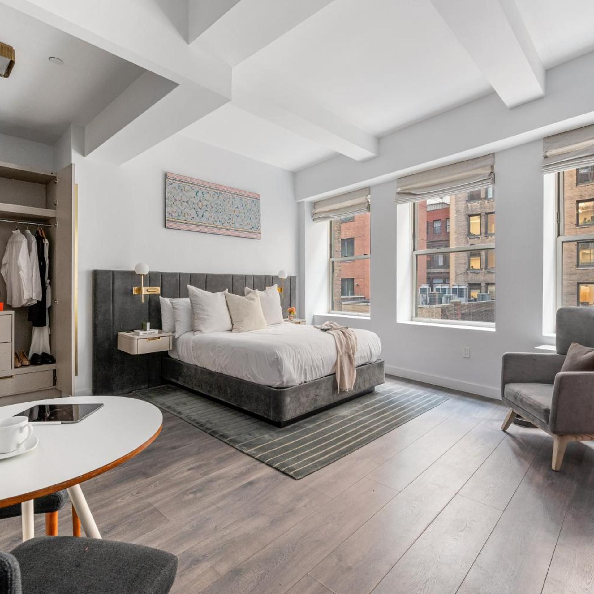 A modern bedroom with a large bed, a bedside table, a chair, and a view of the city through large windows.