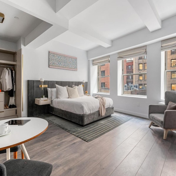 Un dormitorio moderno con una cama grande, una mesita de noche, una silla y vistas a la ciudad a través de amplios ventanales.