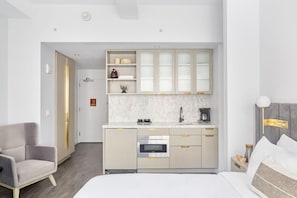 A modern hotel room with a bed, a gray armchair, a kitchenette with a built-in oven, and a small bedside table with a lamp.