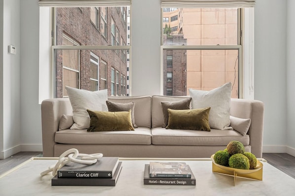 A modern living room with a beige sofa, two windows, and a coffee table with books and a decorative bowl.