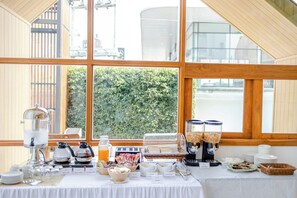 Montaje de un bufé de desayuno con diferentes dispensadores de alimentos y bebidas, una máquina de café y vistas a zonas verdes a través de amplios ventanales.